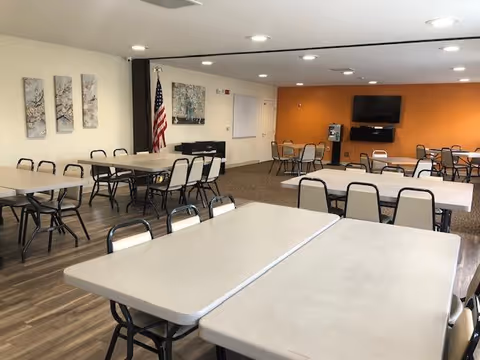 A spacious dining room with multiple rectangular tables and chairs arranged neatly. The room has wood flooring in the foreground and carpet in the background, with an orange accent wall featuring a mounted flat-screen TV and a coffee station. There are three decorative wall art pieces and an American flag on one side of the room.