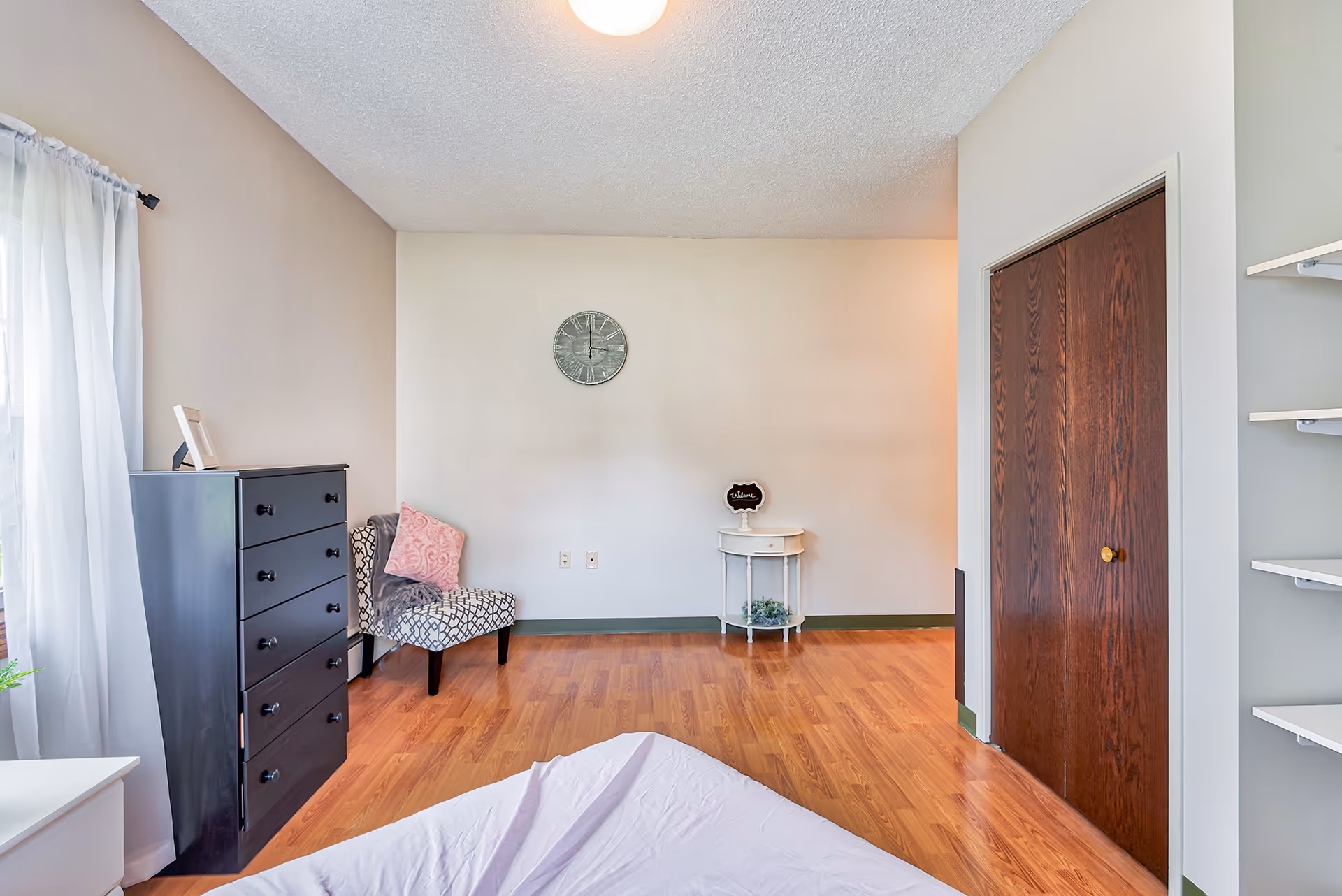 A bedroom with light-colored walls and wood flooring. The room features a black chest of drawers, a patterned chair with a pink pillow and gray throw, a small white round table with a welcome sign, a wall clock, and a wooden closet door. Sheer white curtains cover a window on the left side.