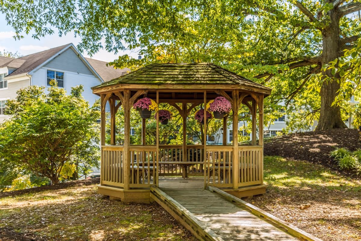 A wooden gazebo with a shingled roof and hanging flower pots, situated in a garden area with trees and bushes. A wooden ramp leads up to the gazebo, and residential buildings are visible in the background.