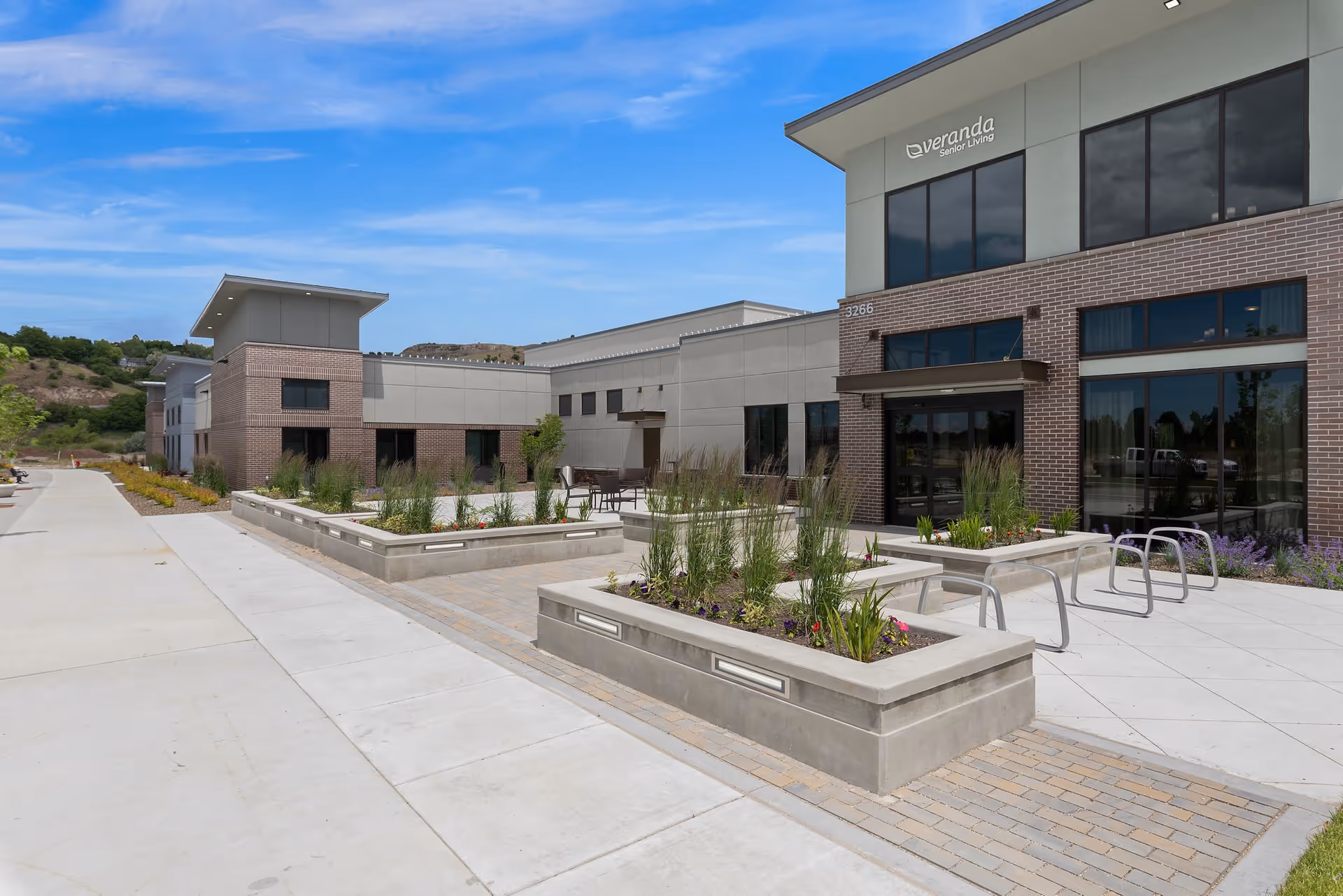 Exterior view of a modern senior living facility with a paved walkway, raised concrete planters with greenery and flowers, and large windows. The building has a combination of brick and light-colored siding, with a clear blue sky above.
