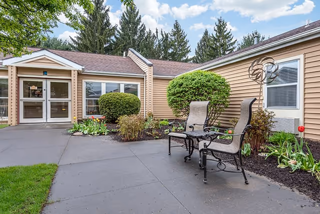 Outdoor patio area at Brookdale Painted Post featuring two cushioned metal chairs with a small table between them, surrounded by landscaped bushes and plants. The building exterior is beige with white trim, and there are large windows and a glass door entrance. Tall evergreen trees are visible in the background under a partly cloudy sky.