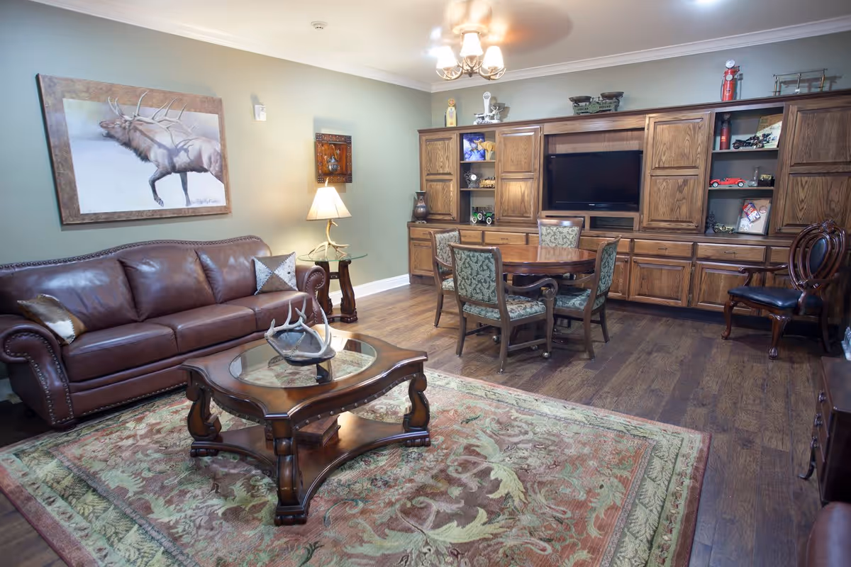 A cozy living room with a brown leather sofa, a wooden coffee table with a decorative antler centerpiece, a patterned area rug, and a wooden dining table with four upholstered chairs. The room features wooden cabinetry with shelves displaying various decorative items and a flat-screen TV. A lamp with a beige shade sits on a small glass-top side table next to the sofa. The walls are painted light green and there is a large framed painting of an elk above the sofa.