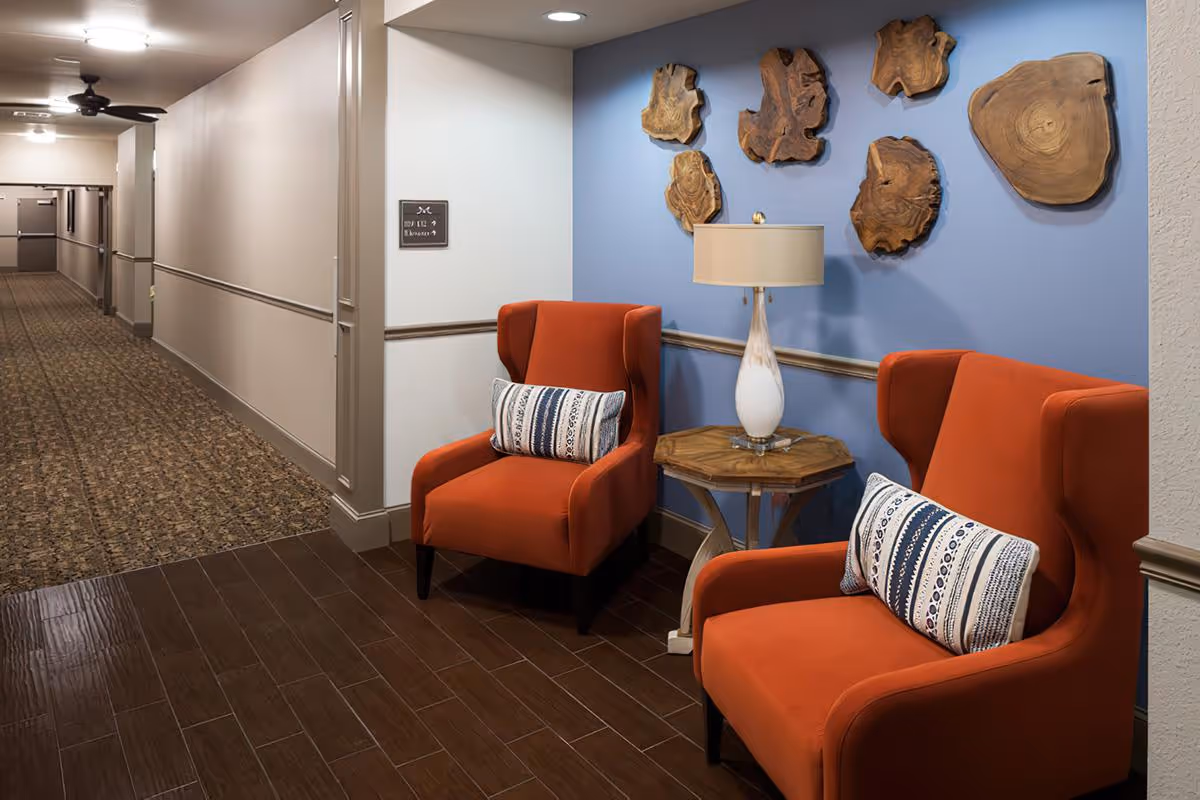 A hallway corner in a senior living facility with two orange armchairs featuring patterned cushions, a small wooden side table with a white lamp, and decorative wooden wall art mounted on a blue accent wall. The hallway has carpeted flooring and beige walls with molding.