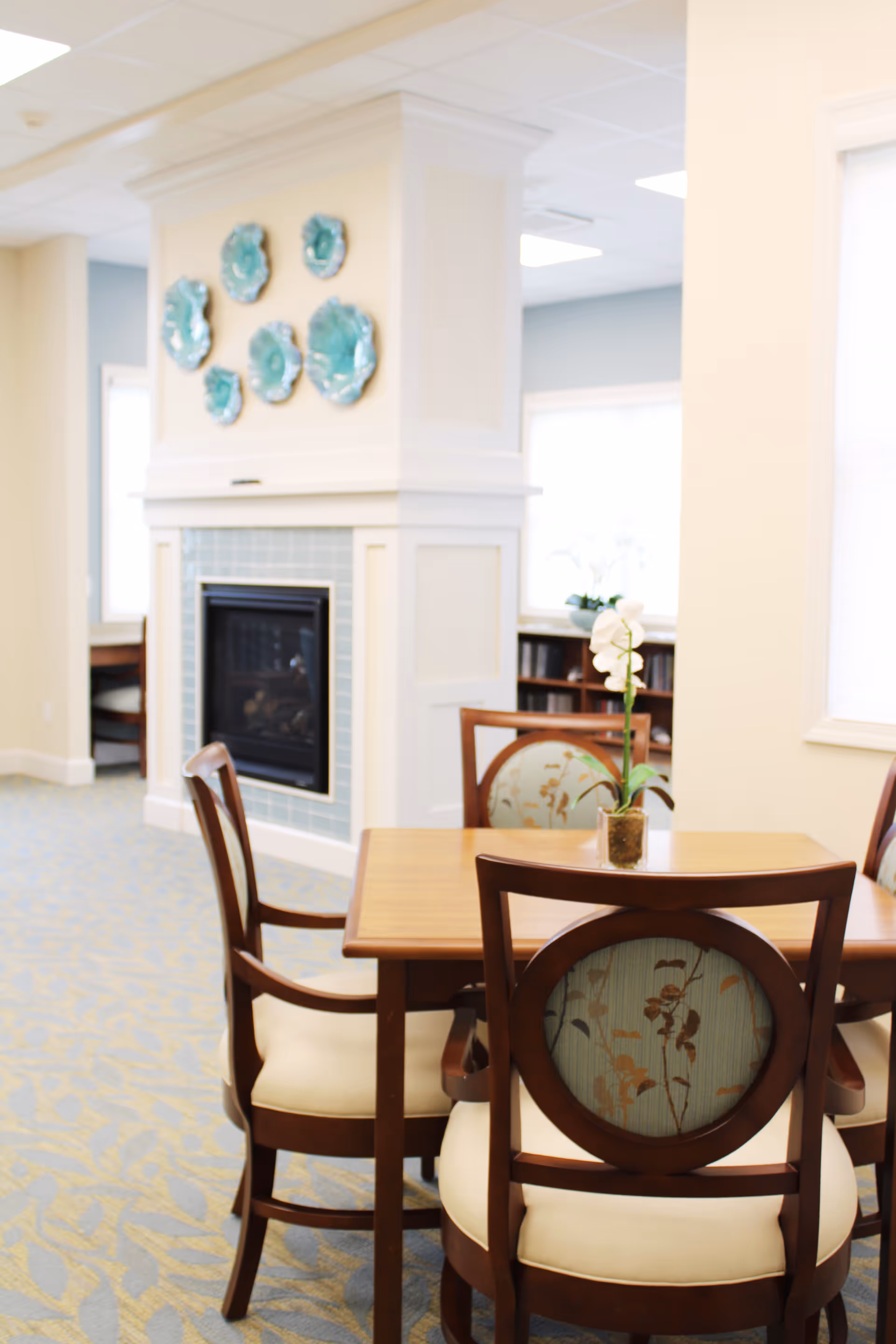 A cozy interior space featuring a wooden table with four upholstered chairs around it. A small potted orchid is placed on the table. In the background, there is a white fireplace with a tiled surround and decorative blue-green ceramic plates mounted above it. The room has soft lighting and carpeted flooring with a subtle pattern.