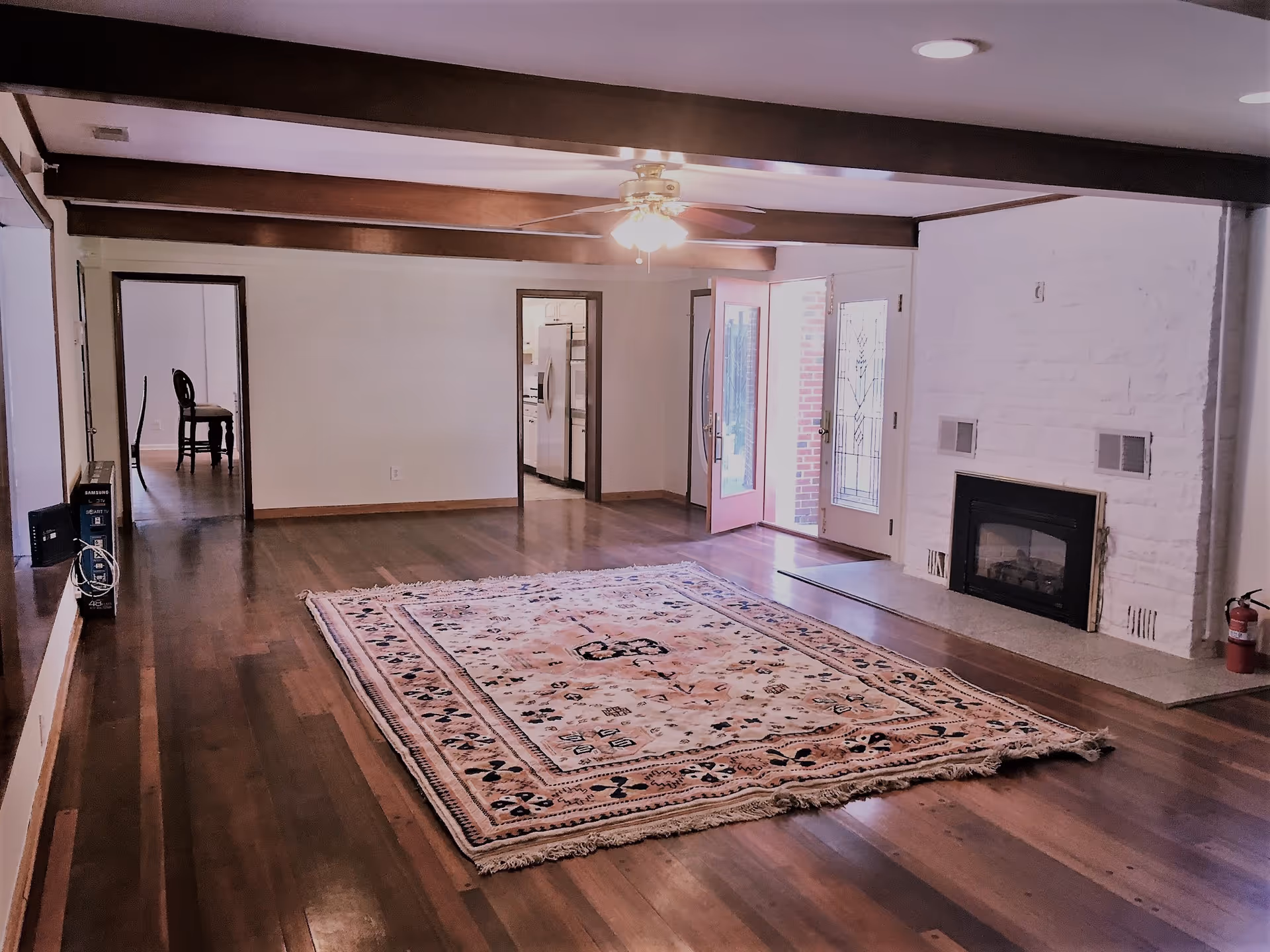 Spacious living room with wooden floors, a large patterned area rug in the center, exposed wooden ceiling beams, a ceiling fan with lights, and a white brick fireplace. There are two doorways leading to other rooms, one showing a kitchen with stainless steel appliances and the other showing a dining area with chairs. A glass door with decorative panes opens to the outside, and a fire extinguisher is placed near the fireplace.