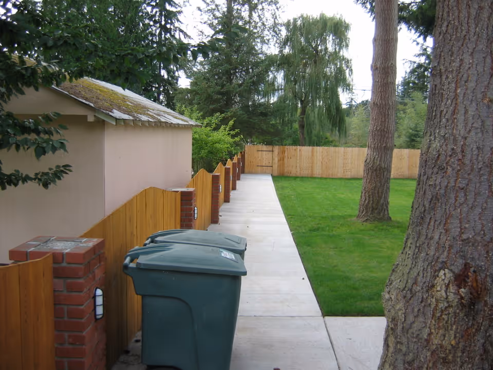Outdoor view of a pathway alongside a wooden fence with brick pillars, green grass lawn, and two large trees. There are two green trash bins next to the fence and a small building with a mossy roof on the left side.