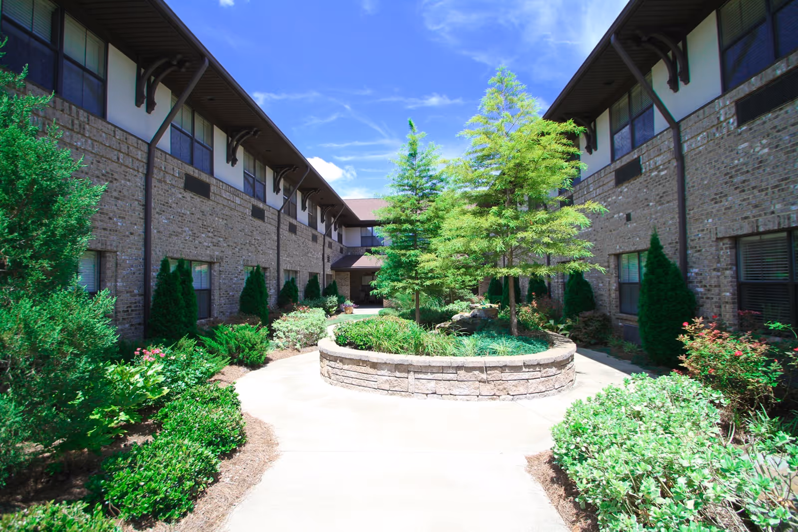 Outdoor courtyard area at Legacy Ridge Trussville featuring a circular raised stone planter with green trees and shrubs, surrounded by a concrete walkway. The courtyard is enclosed by a two-story brick building with windows and greenery along the sides under a bright blue sky with some clouds.