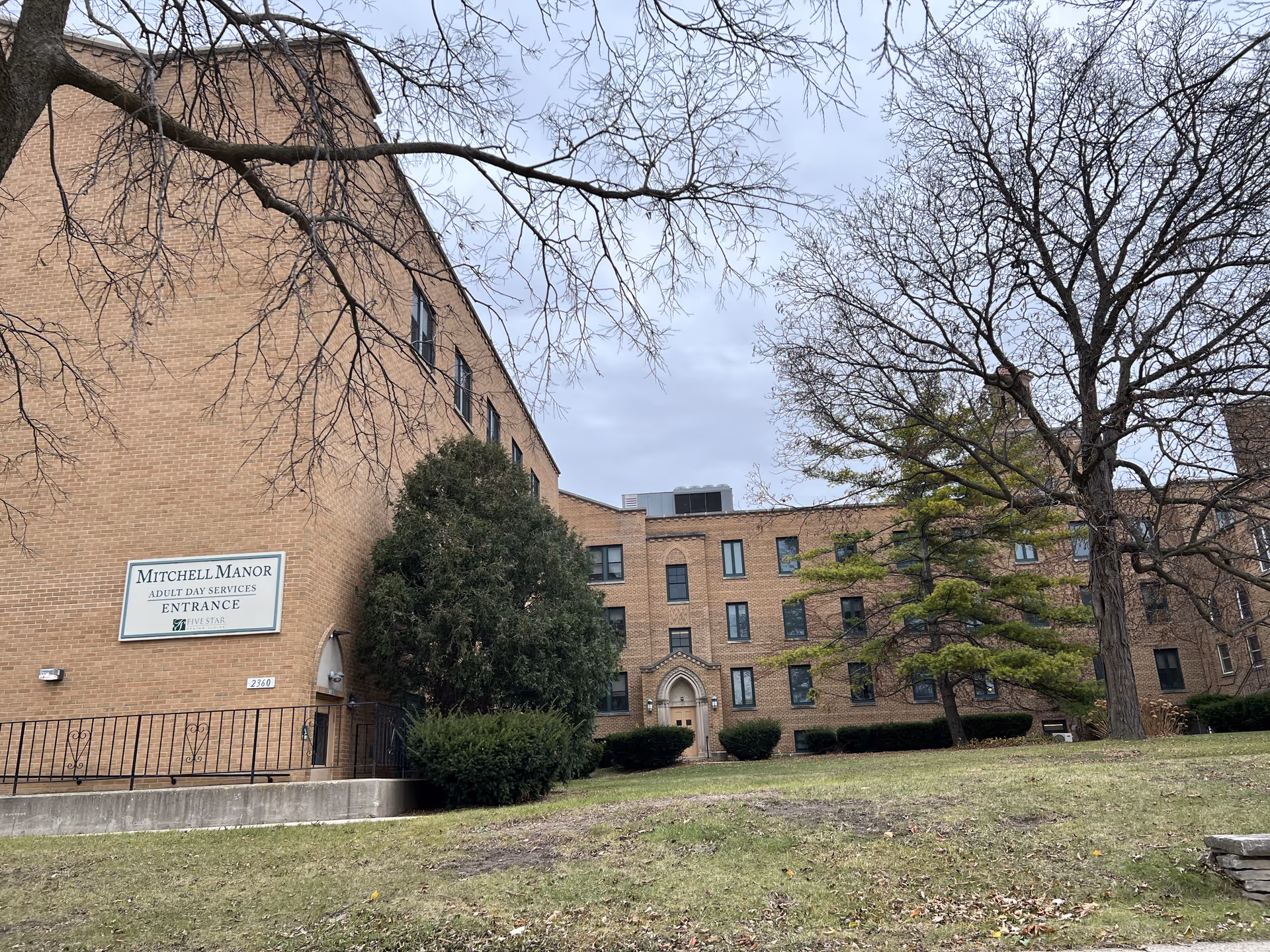 Exterior view of a large brick building with multiple windows and a sign that reads 'Mitchell Manor Adult Day Services Entrance'. The building is surrounded by leafless trees and some green bushes, with a grassy area in the foreground under a cloudy sky.