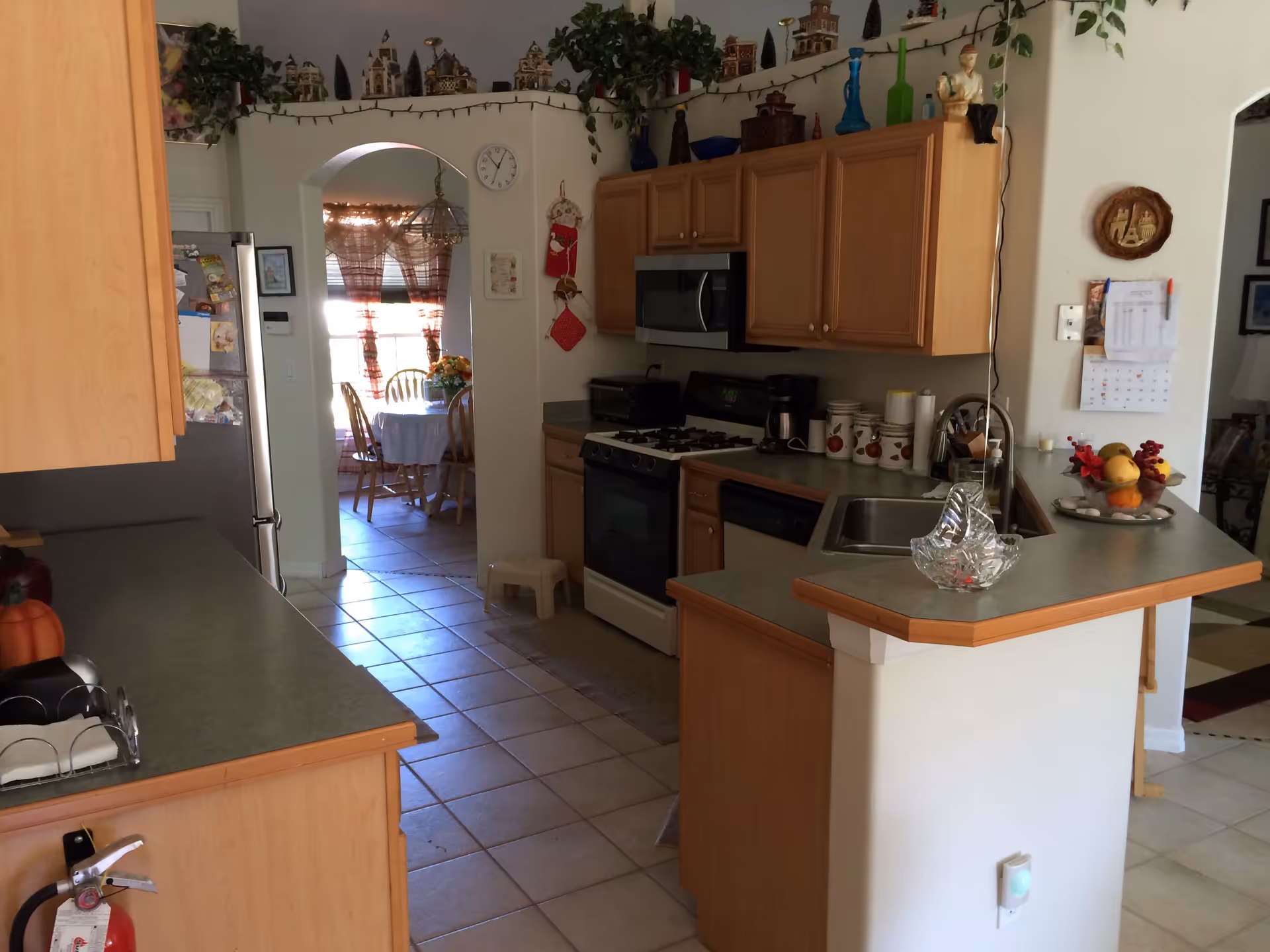 A kitchen with wooden cabinets, a gas stove, microwave, coffee maker, and a sink on a countertop. There is a small stool on the tiled floor and a dining area visible through an arched doorway with a table and chairs near a window with sheer curtains. Decorative items and plants are placed on top of the cabinets.