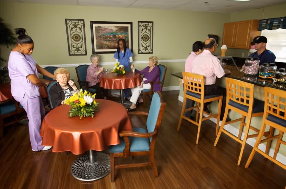 A communal dining area in a senior living facility with elderly residents and caregivers. Two elderly women sit at a round table with a floral centerpiece, one being assisted by a caregiver in purple scrubs. Another table in the background has two elderly women and a caregiver standing beside them. On the right, two elderly men sit on high chairs at a counter where a staff member serves them. The room has wooden flooring, light green walls, and framed artwork on the wall.