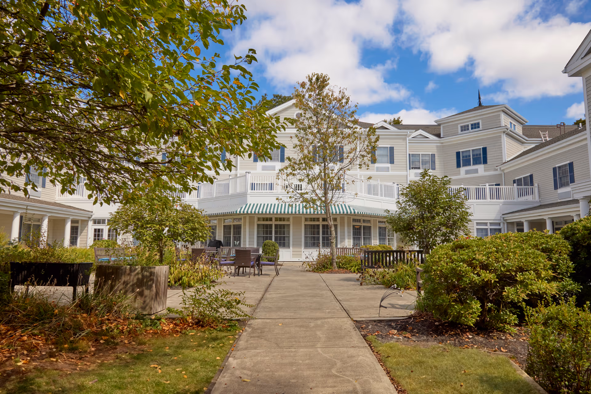 Outdoor courtyard area of Monarch Southbury facility with a paved walkway, green trees, shrubs, benches, and patio tables and chairs. The building is white with multiple windows and a striped green and white awning under a blue sky with some clouds.