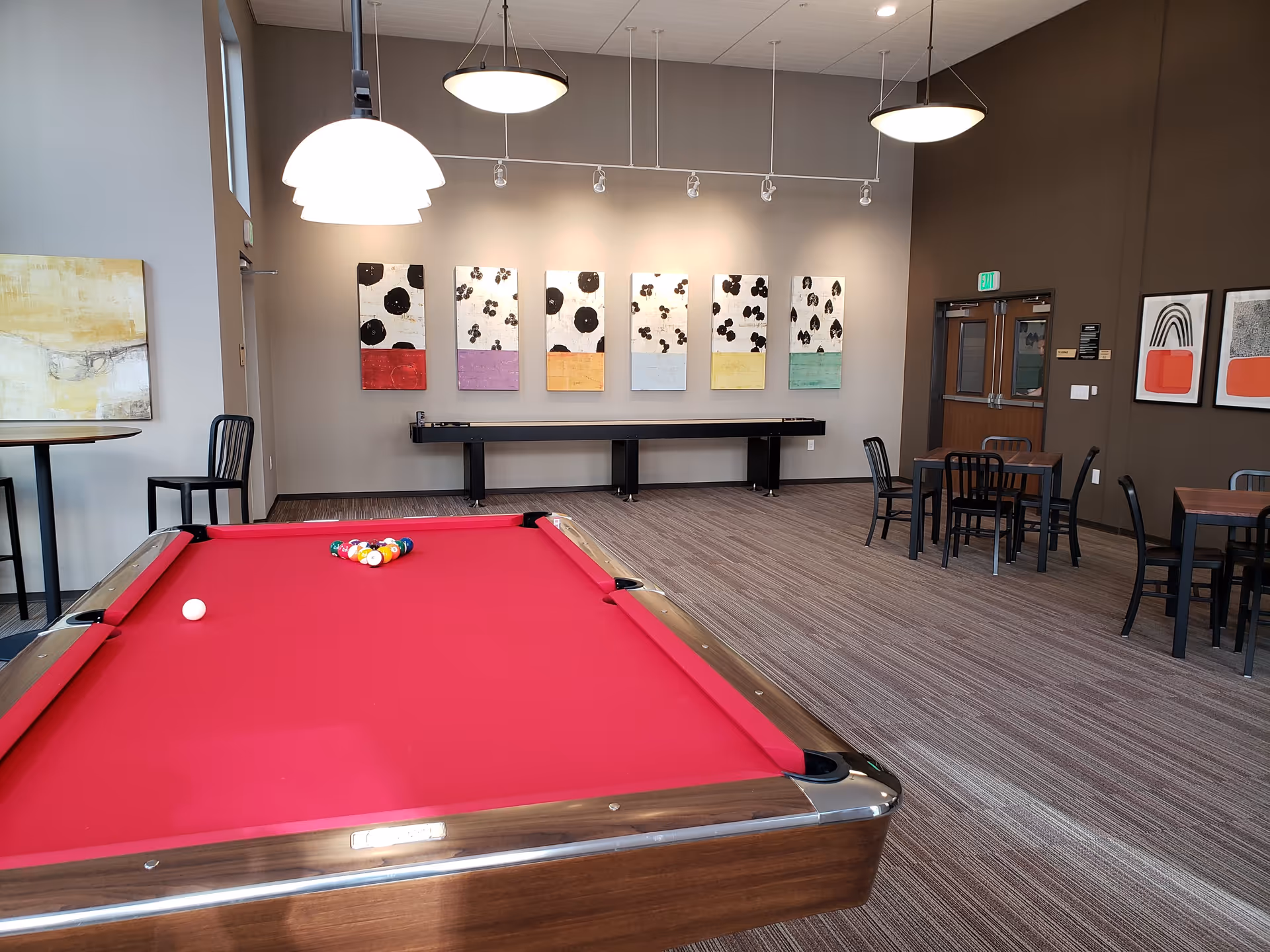 A recreational room with a red felt pool table in the foreground, a shuffleboard table against the far wall, several small tables with black chairs, and modern abstract artwork on the walls. The room has pendant lights hanging from the ceiling and a carpeted floor.