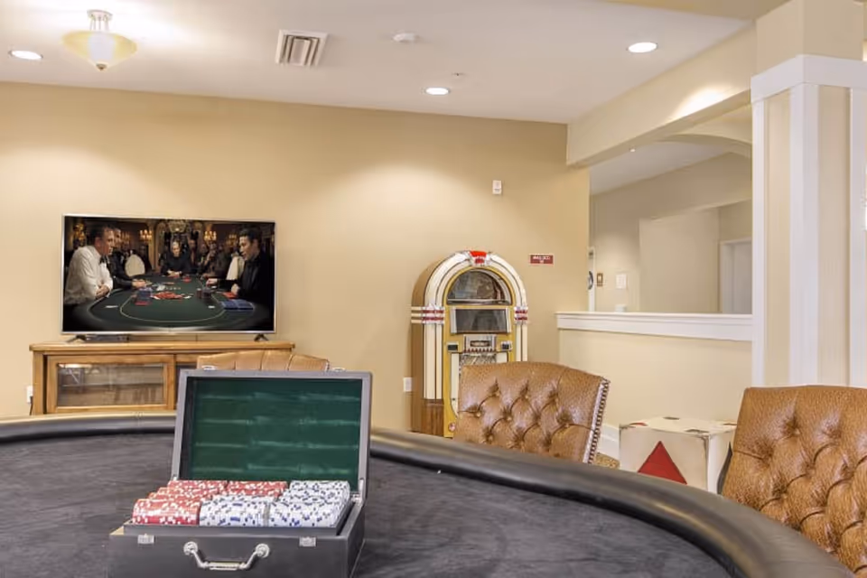 Poker table with a case of chips in a lounge area with leather chairs, a TV showing a card game and a vintage jukebox.