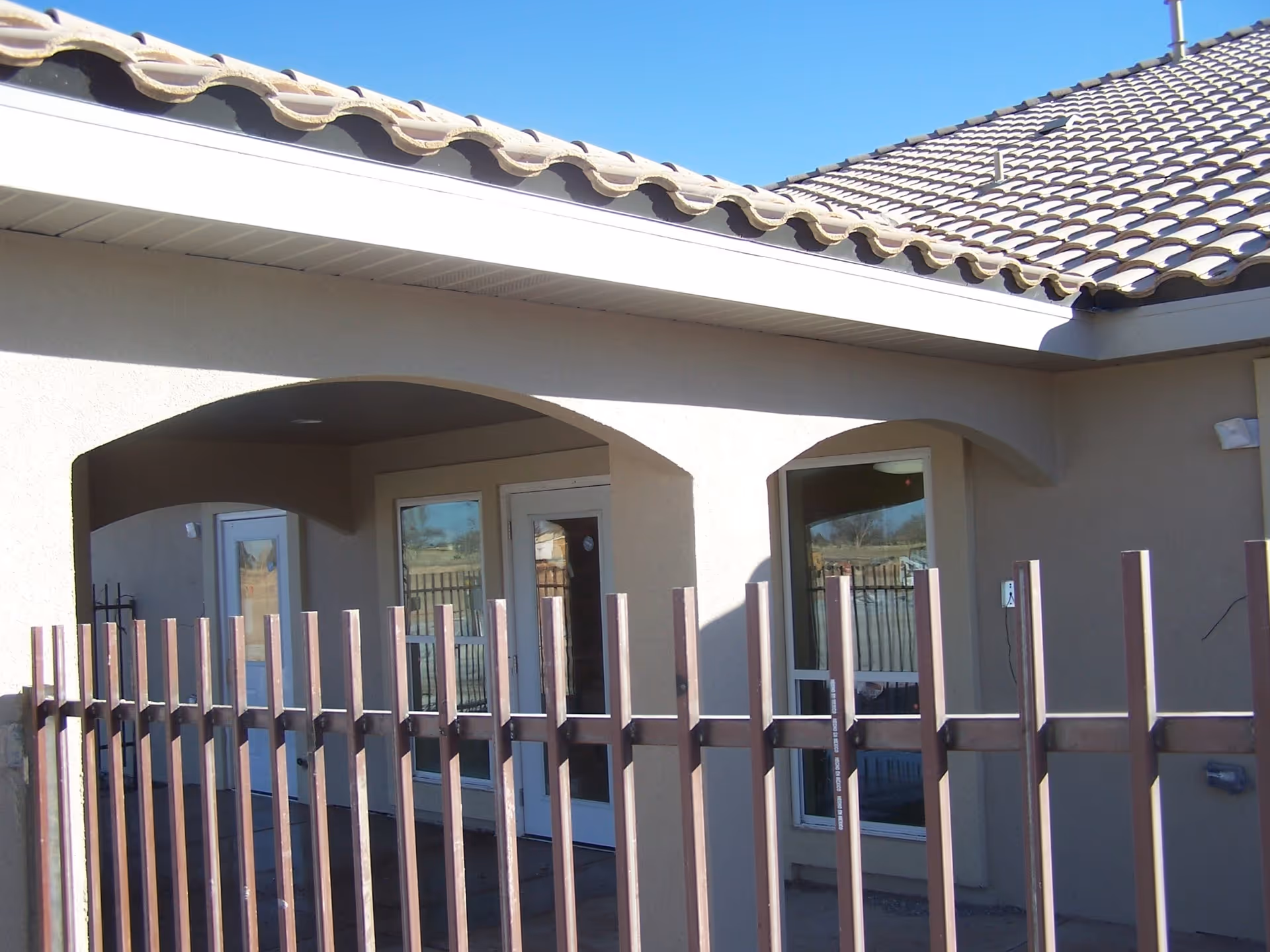 Exterior view of a building with beige stucco walls and a tiled roof, featuring arched doorways and windows behind a brown metal fence under a clear blue sky.
