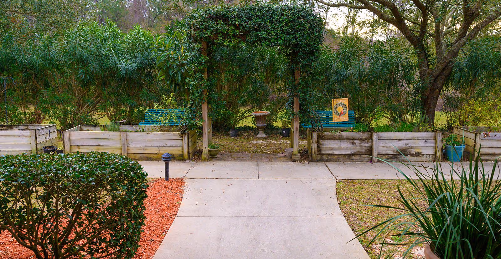A peaceful outdoor garden area with a concrete pathway leading to a wooden arch covered in green vines. On either side of the arch are raised wooden garden beds with various plants. Behind the arch, there are two green benches and a decorative urn. The garden is surrounded by tall green bushes and trees.