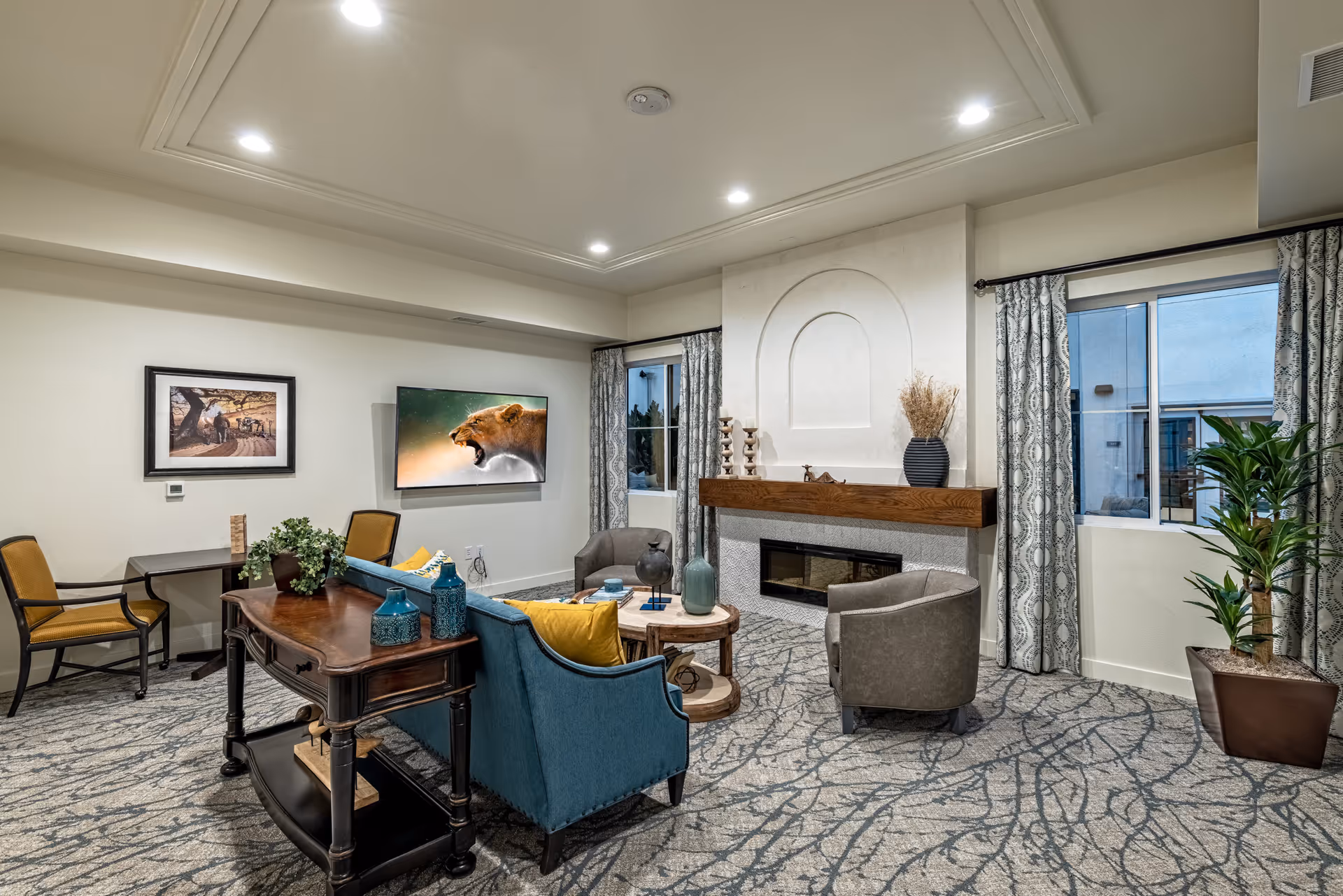 A cozy living room area in Serra Sol Memory Care featuring a blue sofa with yellow pillows, two gray armchairs, a round wooden coffee table, and a wooden console table with decorative vases. The room has a modern fireplace with a wooden mantel, two windows with patterned curtains, a wall-mounted TV displaying a roaring lioness, and a framed picture on the wall. The carpet has a gray and blue abstract pattern, and there is a potted plant near one window.
