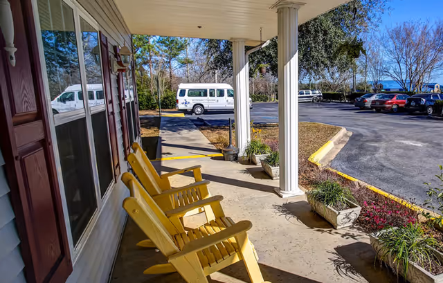 Covered front porch with three yellow wooden chairs overlooking a parking lot and a white facility van.