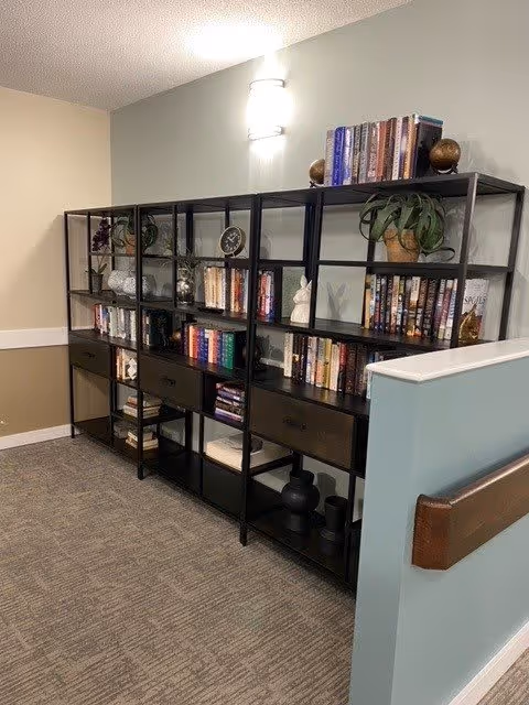 A hallway corner in a senior living facility with a black metal bookshelf filled with books, decorative items, and plants. The walls are painted in light beige and light blue, with a wall-mounted light fixture above the bookshelf. The floor is carpeted in a neutral tone.