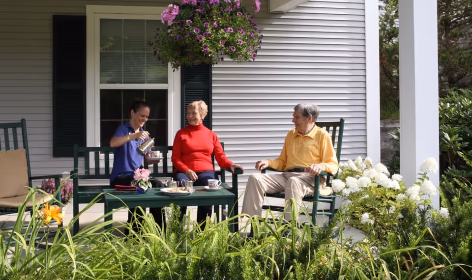 Three people sitting on a porch outside a house, with one person pouring tea or coffee into a cup. They are seated on green chairs around a green table with a tray of cookies and cups. There are flowers and greenery around the porch, including hanging purple flowers and white hydrangeas.