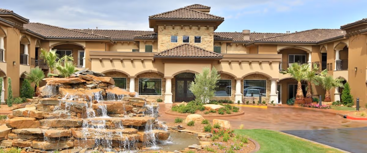 Exterior view of The Retreat at Sunbrook facility featuring a large stone waterfall fountain in the foreground, surrounded by landscaped greenery and palm trees, with a two-story building with tiled roof and arched windows in the background.