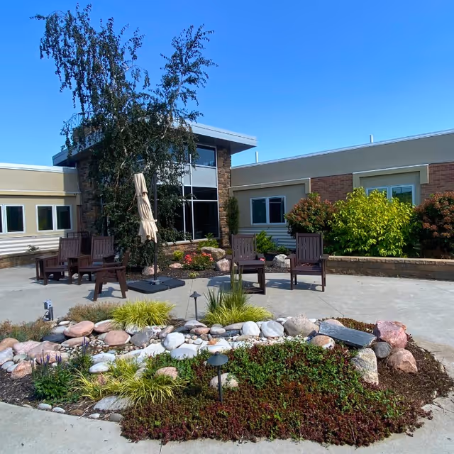 Outdoor patio area at Memorial Community Health with several wooden chairs arranged around a closed beige umbrella. The patio is surrounded by landscaped garden beds with rocks, green shrubs, and small plants. The building exterior features large windows, stone accents, and beige siding under a clear blue sky.