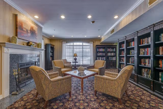 A cozy living room with four upholstered armchairs arranged around a wooden coffee table on a patterned carpet. The room features a fireplace with a mantel decorated with small sculptures and a painting above it. Large windows with blinds and a table lamp provide natural and artificial light. Dark wooden bookshelves filled with books line the walls.