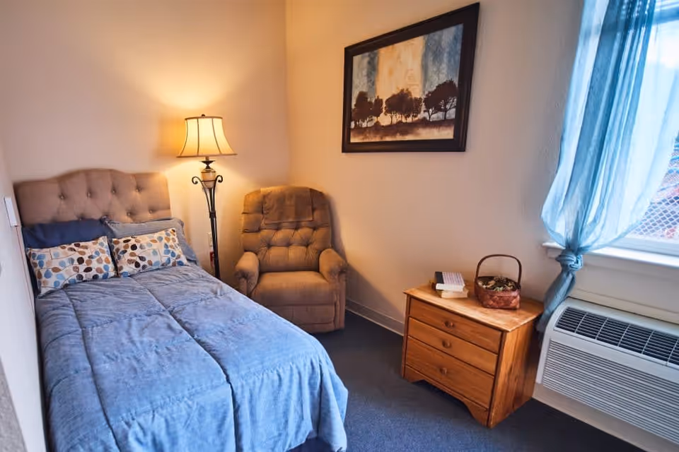 A cozy bedroom with a single bed covered in a blue comforter and patterned pillows. Next to the bed is a tall floor lamp and a cushioned armchair. A wooden nightstand with three drawers holds a basket and some books. A window with sheer blue curtains lets in natural light, and a framed painting of trees hangs on the wall above the armchair.