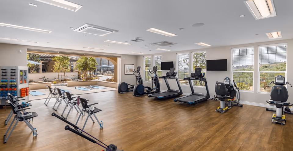 A spacious senior living facility fitness room with wooden flooring, several exercise machines including treadmills, stationary bikes, and elliptical trainers. There are also folding chairs arranged in rows and a rack of colorful dumbbells on the left side. Large windows and an open sliding door provide natural light and a view of an outdoor patio area with plants and seating.