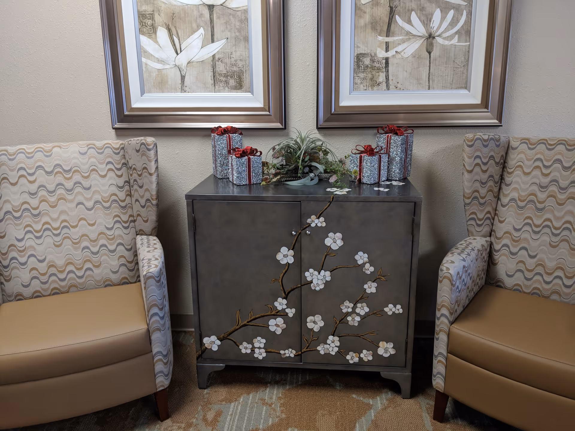 Two patterned armchairs flank a decorative cabinet with a floral design topped with small gift boxes and a plant, and framed botanical prints hang above.