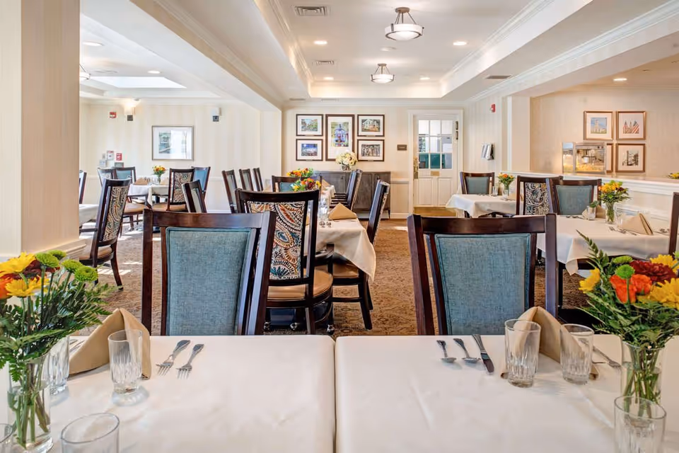 A bright and elegant dining room with tables covered in white tablecloths, set with glasses, silverware, and beige napkins. The chairs have wooden frames with blue and patterned upholstery. Fresh flower arrangements decorate the tables. The room has framed pictures on the walls, recessed lighting, and a carpeted floor.