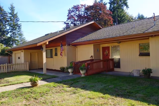 A single-story beige house-style facility with a front porch, railing, flag, and greenery.