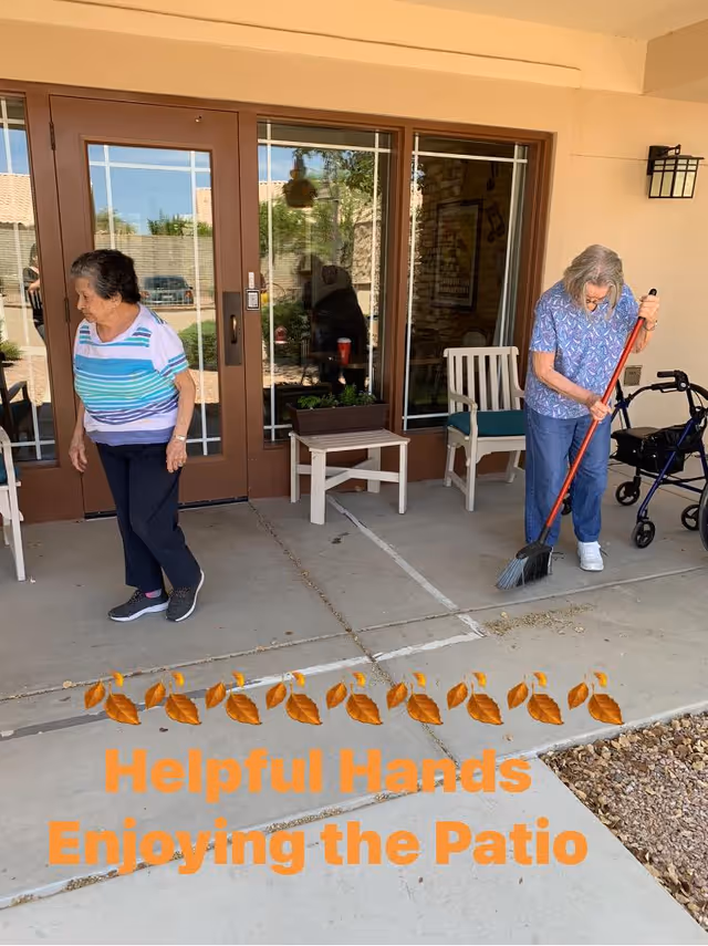 Two elderly women on a patio outside a building. One woman is sweeping the ground with a broom, while the other woman stands nearby. There are chairs and a small table with a planter on the patio. A walker is visible next to the woman sweeping.