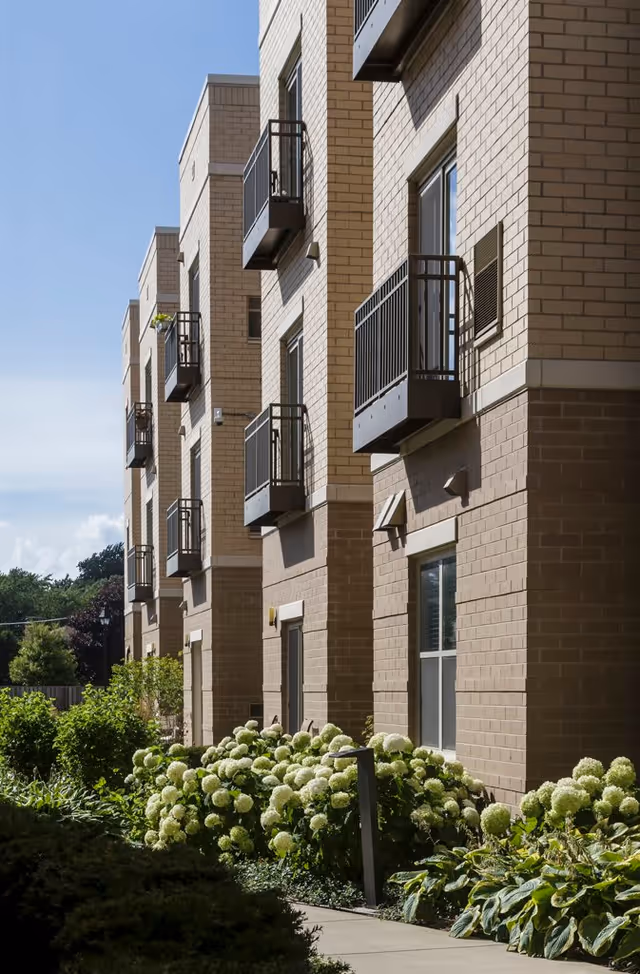 Exterior view of a multi-story brick building with small balconies on each floor. The building is surrounded by well-maintained landscaping including green shrubs and white flowering plants under a clear blue sky.