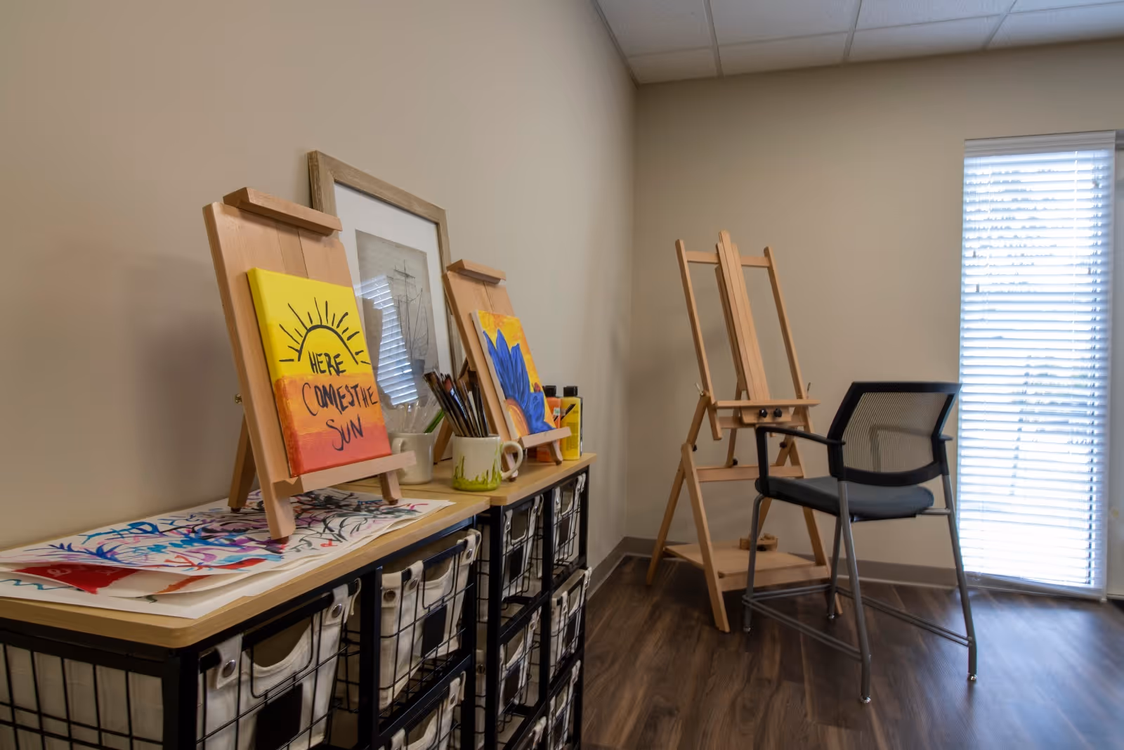 A small art studio corner with two wooden easels, one holding a painting with a sun and the words 'Here Comes The Sun'. There are paintbrushes in a mug, art supplies, and storage bins on a black metal and wood shelving unit. A black mesh chair is positioned near a window with blinds, and the room has wood flooring and beige walls.