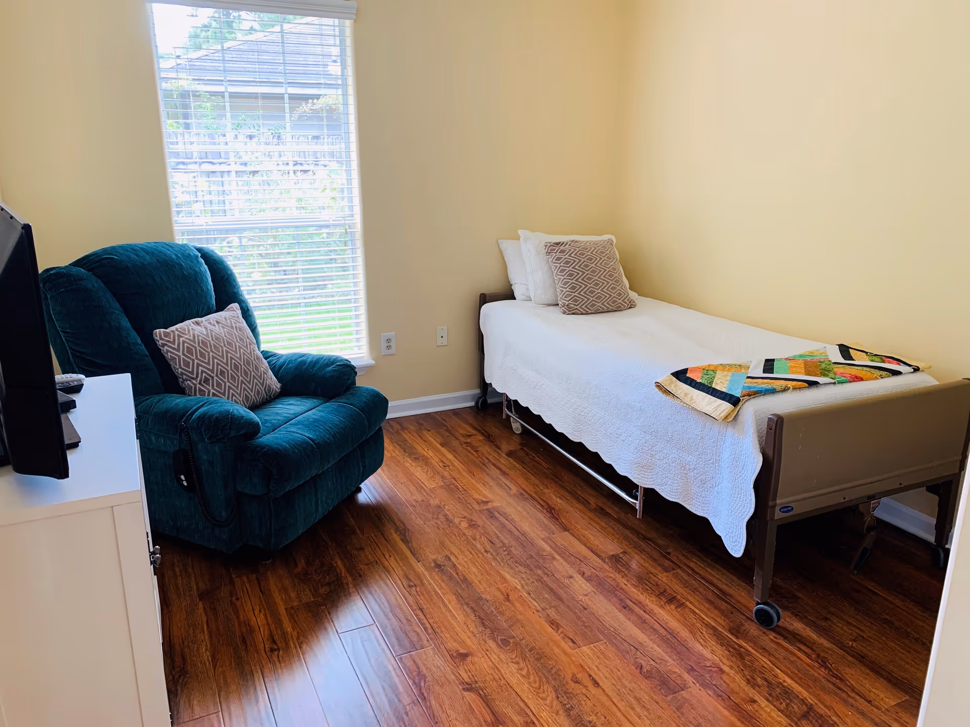 A small, cozy bedroom with a single bed covered in a white quilt and a colorful patchwork blanket at the foot. There are two pillows on the bed. Next to the bed is a green upholstered recliner chair with a patterned cushion. A window with blinds lets in natural light, and a TV is placed on a white stand opposite the chair. The room has wooden flooring and light yellow walls.