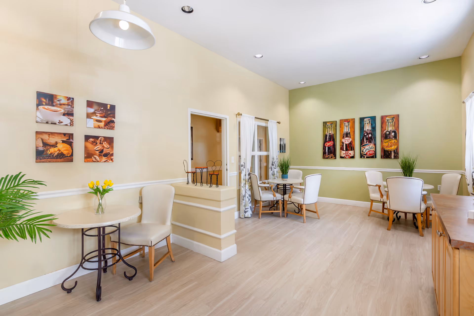 A bright and clean common area with light wood flooring and cream and green walls. There are several small round tables with cream-colored chairs around them. The walls are decorated with colorful paintings of coffee and soda bottles. A vase with yellow tulips is on one table, and potted plants are placed on other tables. The room has recessed lighting and a large hanging light fixture.