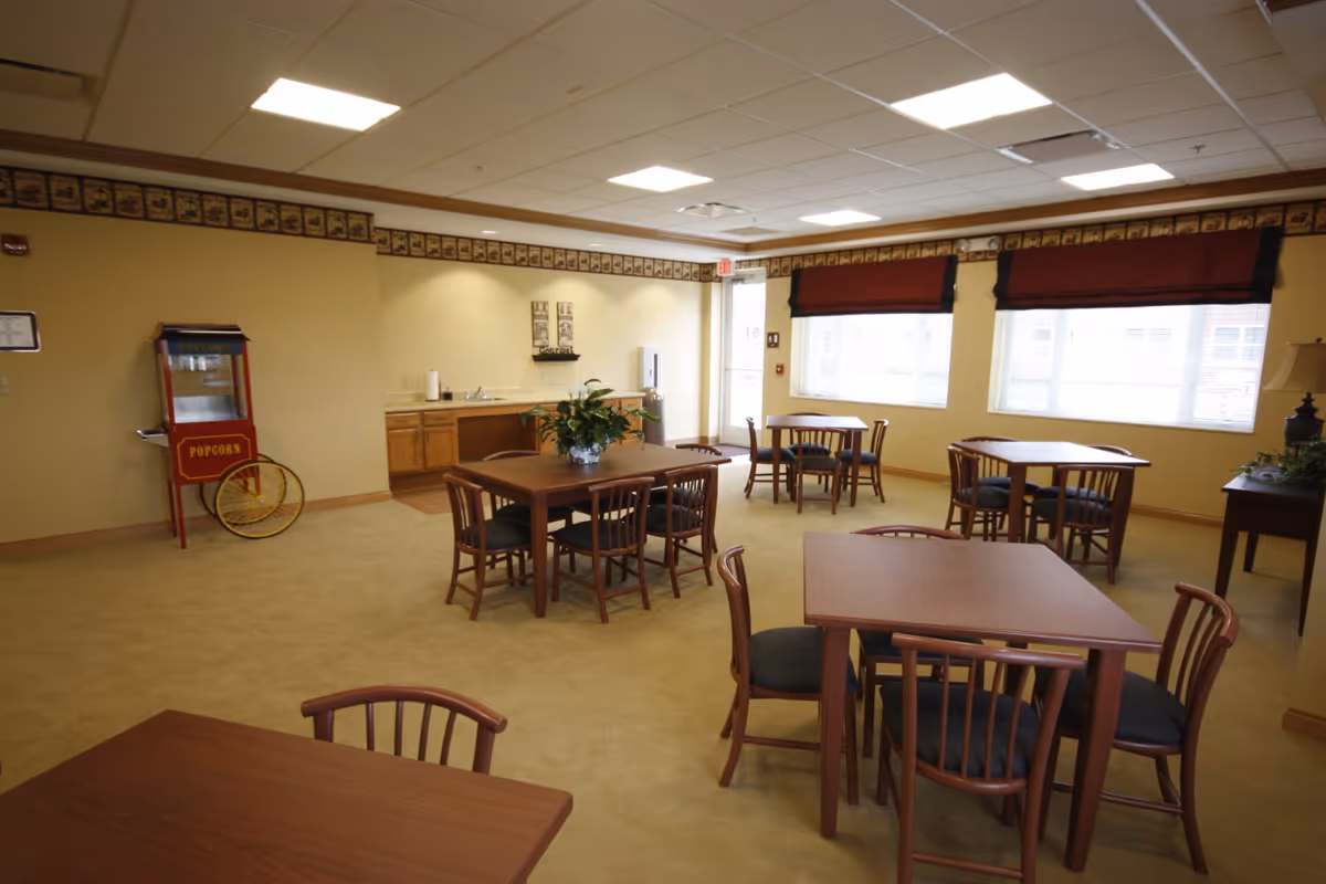 A well-lit dining area with several wooden tables and chairs arranged neatly. There is a popcorn machine against one wall, a small kitchenette with a sink and cabinets, and large windows with red valances letting in natural light. The room has beige walls and carpet, with decorative border trim near the ceiling.