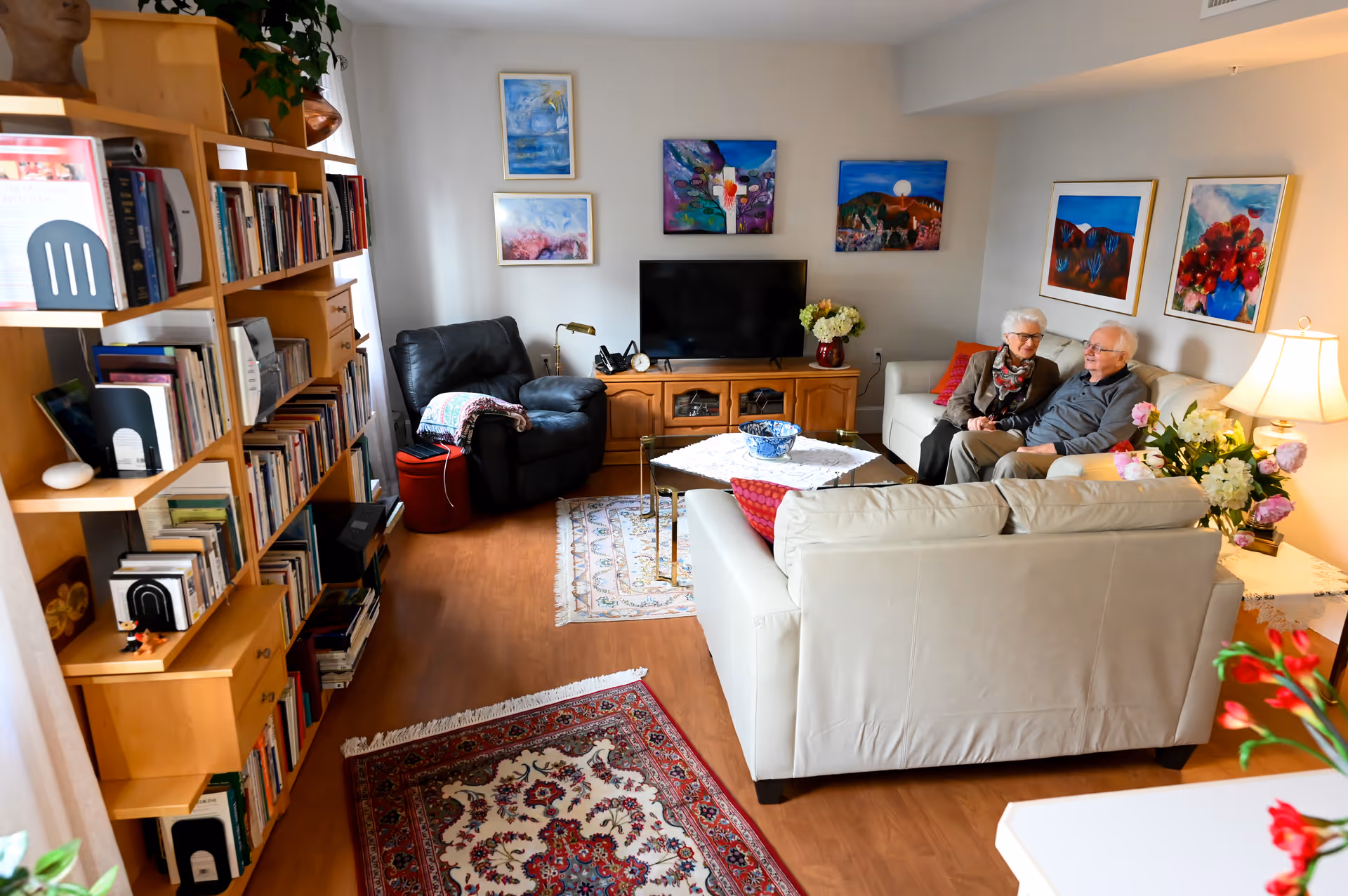 A cozy living room with wooden flooring, a large bookshelf filled with books on the left, a black recliner chair, a wooden TV stand with a flat-screen TV, and two white sofas. An elderly couple is sitting on one of the sofas, smiling and conversing. The walls are decorated with colorful paintings, and there are flowers on the coffee table and side table with a lamp.