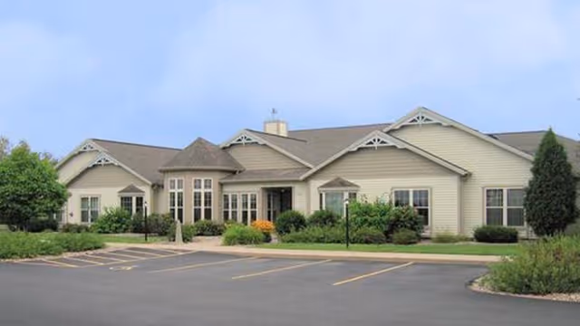 Exterior view of a single-story assisted living facility building with beige siding, multiple windows, and a landscaped front area with bushes and trees under a clear blue sky. There is an empty parking lot in front of the building.