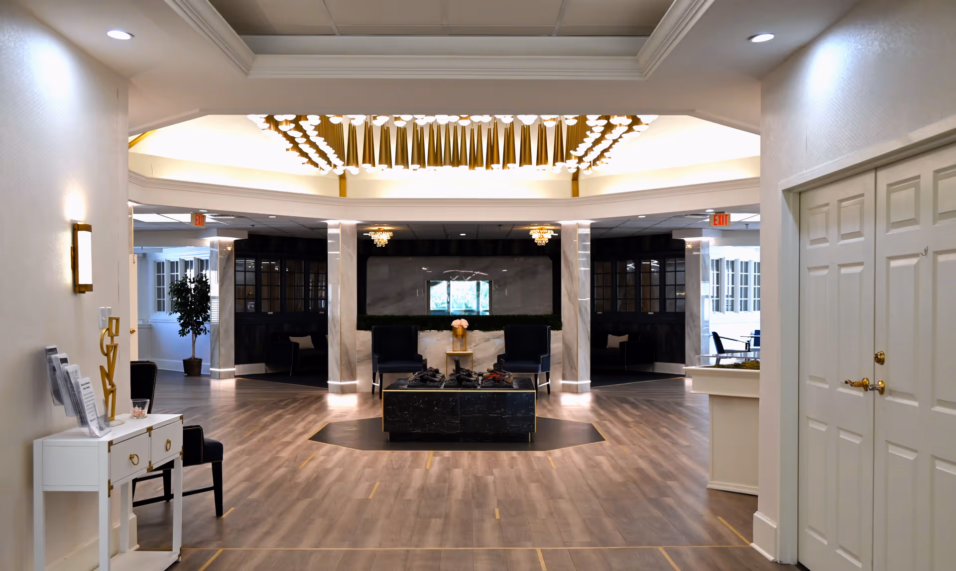 Spacious and elegant interior common area with wood flooring, a central seating arrangement featuring two dark chairs and a low table with decorative items, illuminated by a modern chandelier with gold accents. The area is surrounded by white walls, columns, and large windows allowing natural light, with exit signs visible in the background.