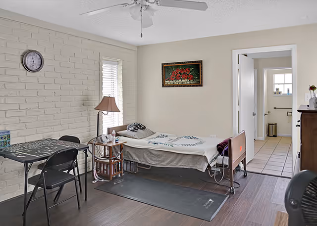 Furnished memory-care bedroom with a single bed, small table and chairs, lamp, and an open doorway to a bathroom.