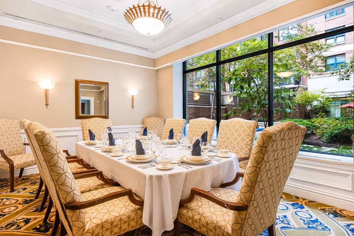 A formal dining room with a rectangular table covered with a white tablecloth, set with plates, cups, glasses, and silverware. Eight upholstered chairs with patterned fabric surround the table. Large windows provide a view of a garden area outside. The room has beige walls, two wall sconces, a mirror, and a decorative ceiling light fixture.