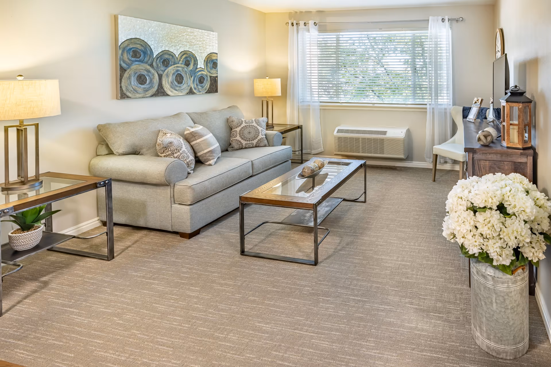 A bright and cozy living room with a light gray sofa adorned with patterned pillows, a glass-top coffee table with decorative items, two side tables with lamps, a large window with white curtains, a wooden console table with a TV and decorative pieces, and a large vase filled with white flowers on the carpeted floor.
