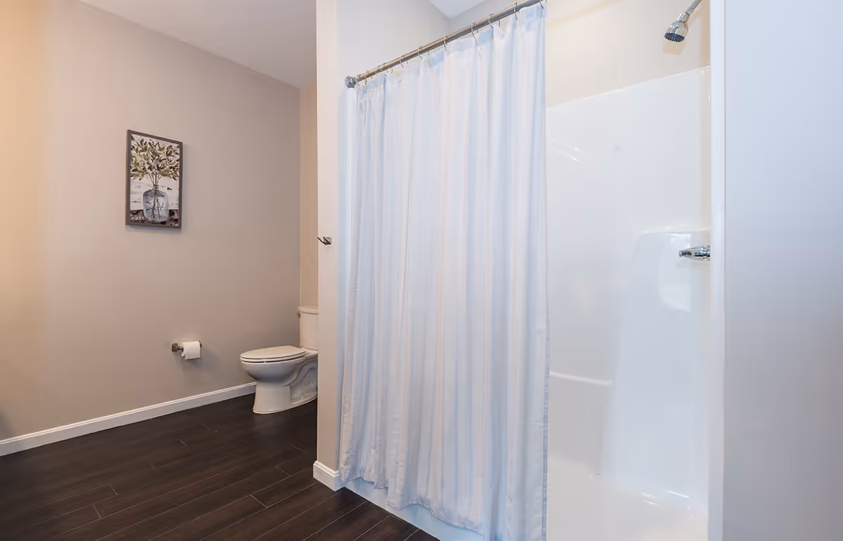 A bathroom with a white shower area covered by a white shower curtain, a toilet with a toilet paper holder on the wall, dark wood flooring, and a framed picture of a plant on the beige wall.