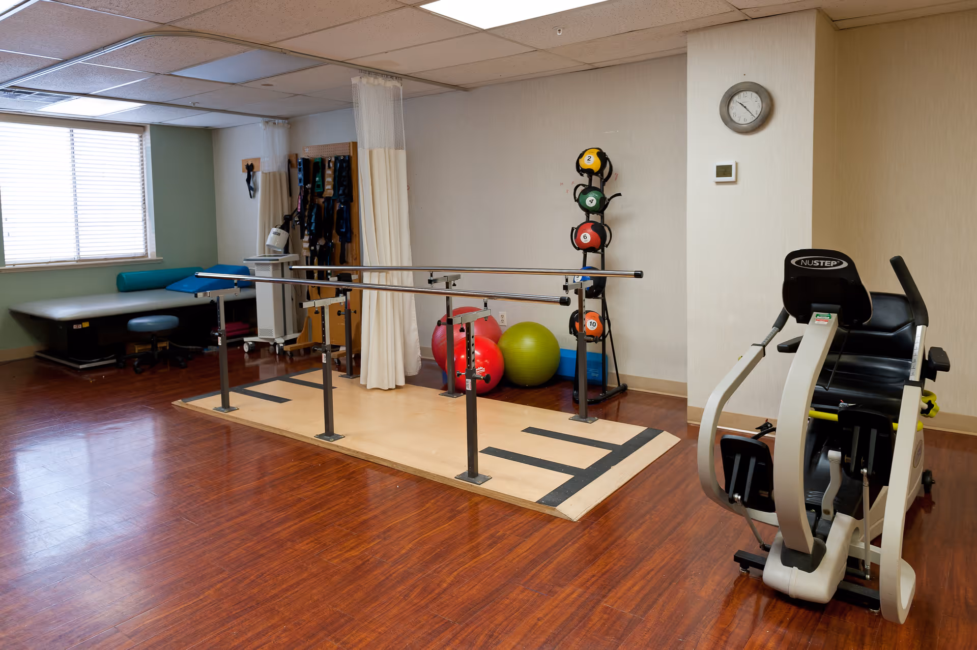 A physical therapy or rehabilitation room with parallel bars for walking exercises, exercise balls, a rack of colorful medicine balls, a stationary exercise machine, and a padded examination or treatment table near a window with blinds.