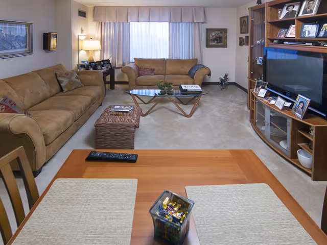 A cozy living room with two beige sofas, a glass coffee table, an entertainment center with TV and framed photos, and a dining table in the foreground.