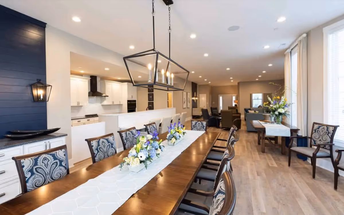 Spacious open-plan dining area with a long wooden table, floral centerpieces, upholstered chairs, and views into the kitchen and seating area.
