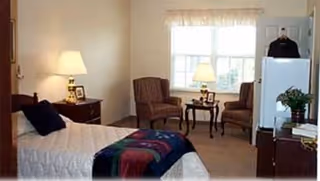A cozy bedroom in a senior living facility featuring a single bed with a colorful blanket, two upholstered chairs around a small table with a lamp, a window with sheer curtains, a nightstand with a lamp and picture frame, and a small refrigerator with a plant on top.