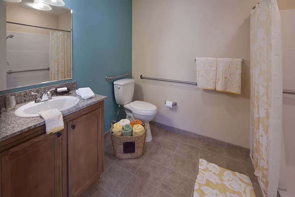 A clean and well-lit bathroom with a wooden vanity topped with a granite countertop and a white sink. Above the sink is a large mirror with three light fixtures. To the right, there is a white toilet with a basket of rolled towels in various colors placed on the floor beside it. The walls are painted in beige and teal, and there are grab bars installed near the toilet and along the wall. A shower with a floral-patterned curtain and matching bath mat is visible on the right side. Two folded towels hang on a towel rack on the beige wall.