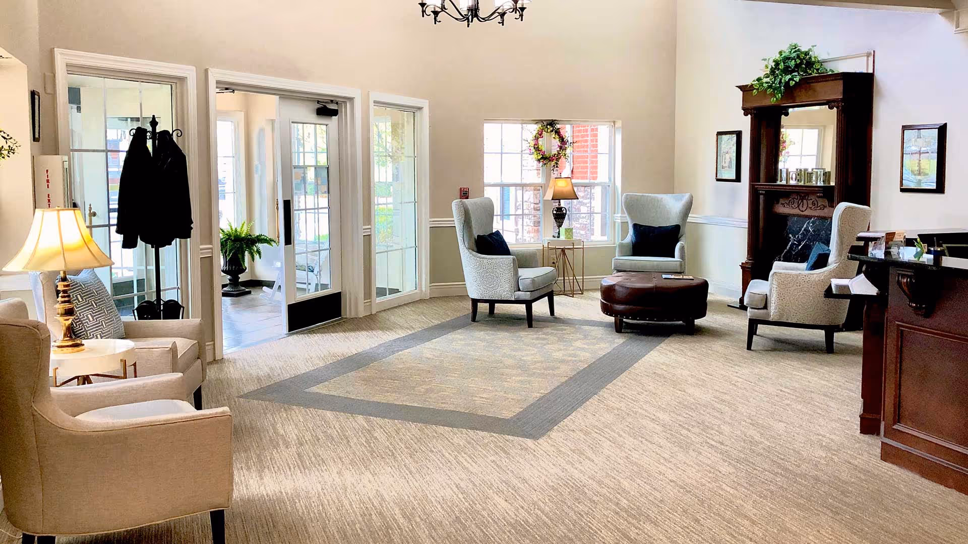 A bright and welcoming living room area with beige carpet and light-colored walls. The room features several upholstered armchairs arranged around a round brown ottoman. There is a wooden fireplace mantel with decorative items and a green plant on top. A table lamp is placed on a small side table next to one of the chairs. Large windows allow natural light to fill the space, and a coat rack with jackets is near the glass doors leading outside.
